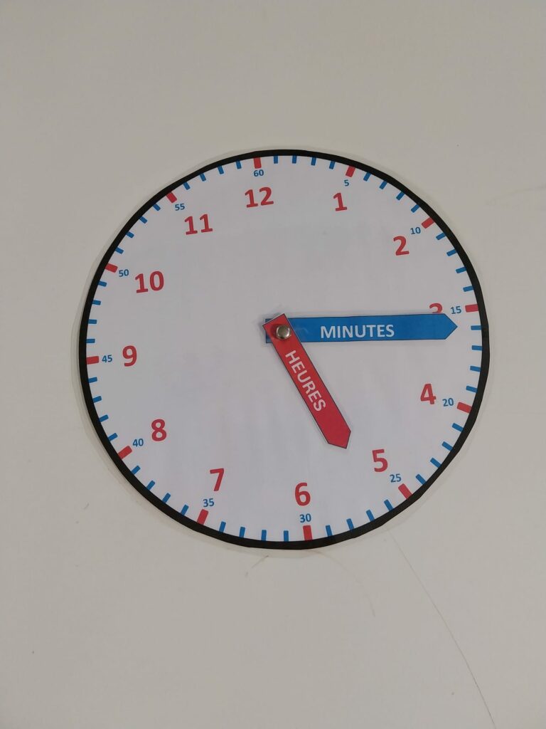 Horloge pédagogique construite et assemblée avec attache parisienne, aiguilles rouges et bleues indiquant heures et minutes.